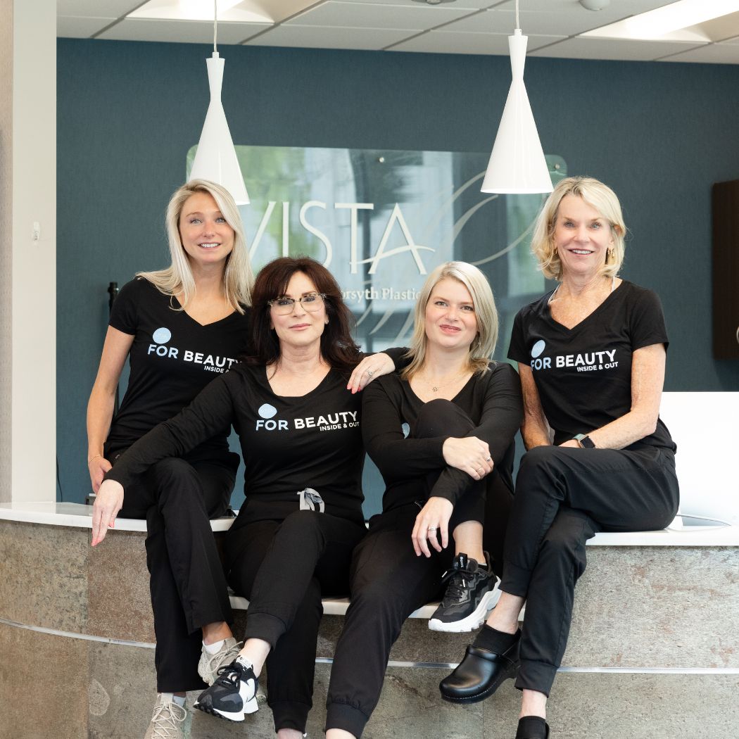 The VISTA Med Spa team sit together on the front desk ledge inside the practice, smiling and wearing matching black "FOR BEAUTY" shirts. The modern reception area with the VISTA logo is visible in the background.