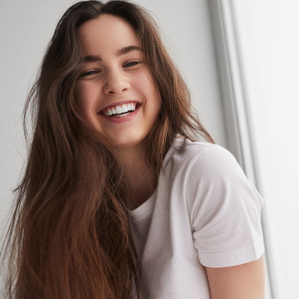 A young woman with long, natural brown hair smiles brightly while sitting near a window in casual clothing. She looks healthy and radiant, conveying happiness and confidence.