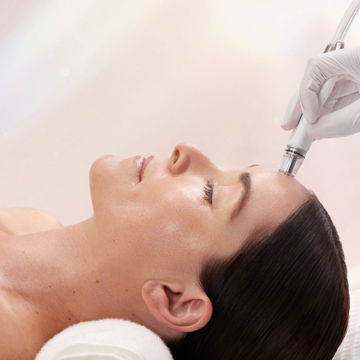 A woman reclines with eyes closed while a clinician uses a DiamondGlow® device on her forehead. The treatment head is applied gently to the skin, highlighting a relaxing, in-progress skincare service.