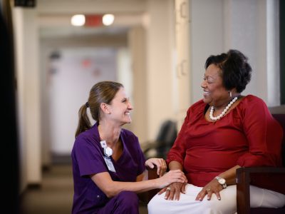 A smiling healthcare professional in purple scrubs kneels beside a seated woman wearing a red blouse and pearl necklace. They are holding hands and sharing a warm conversation in a bright, welcoming hallway.